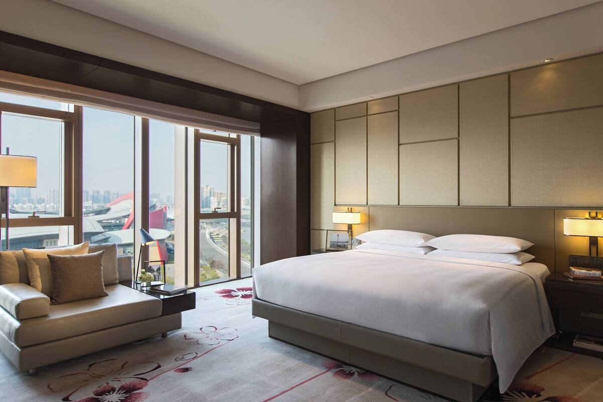 A modern hotel room with a large bed, a sofa, and a view of the city through large windows.