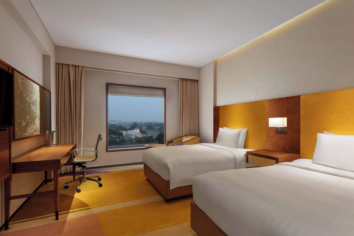 A hotel room with two beds, a desk, a chair, a nightstand, and a window with a view.