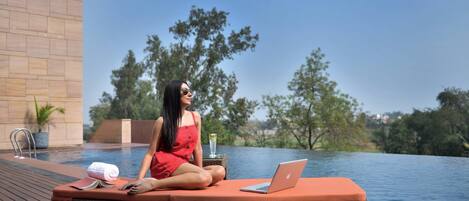 A woman sitting on a wicker lounge chair by a pool with a laptop and a book.