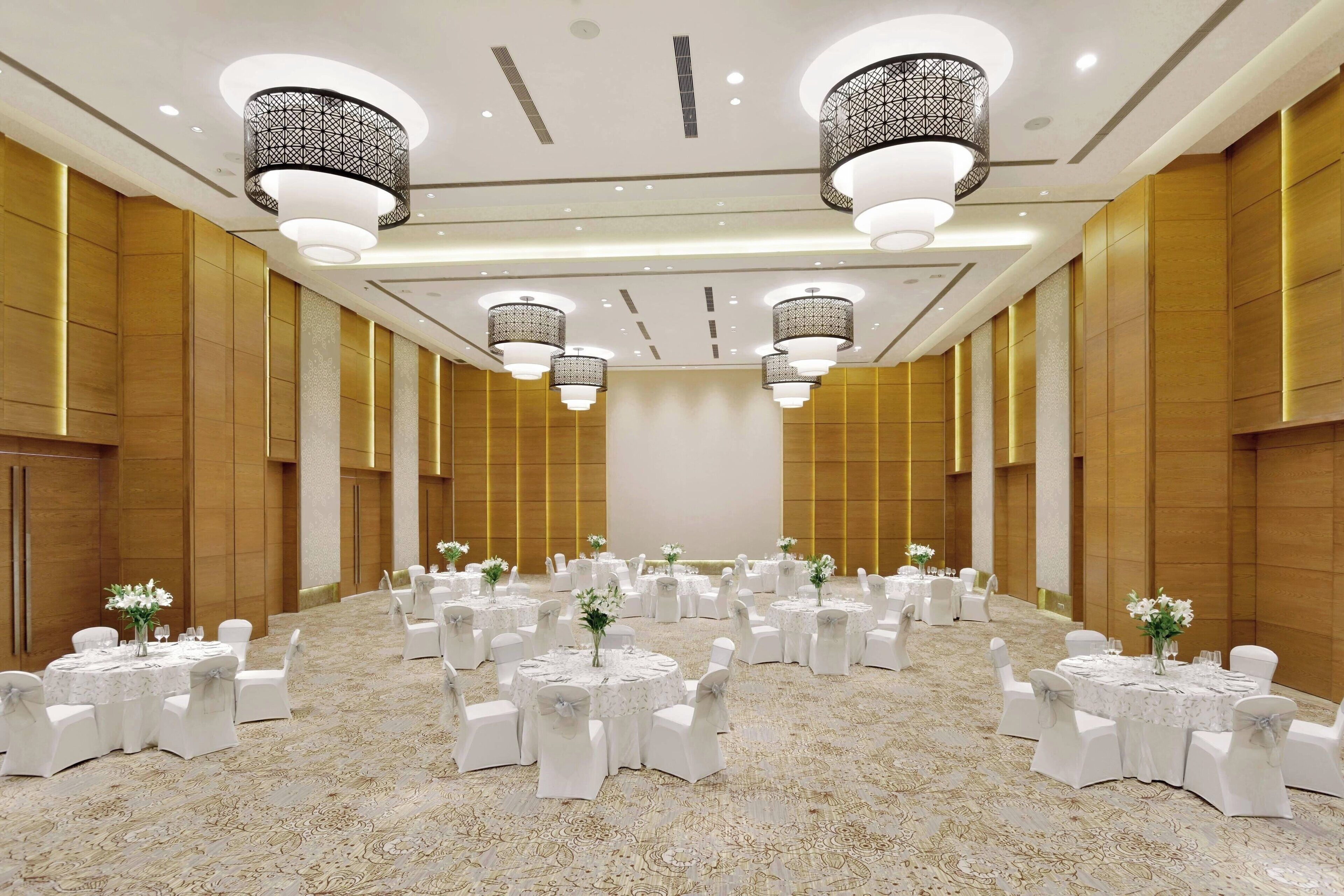 A spacious banquet hall with round tables, white chairs, and chandeliers.