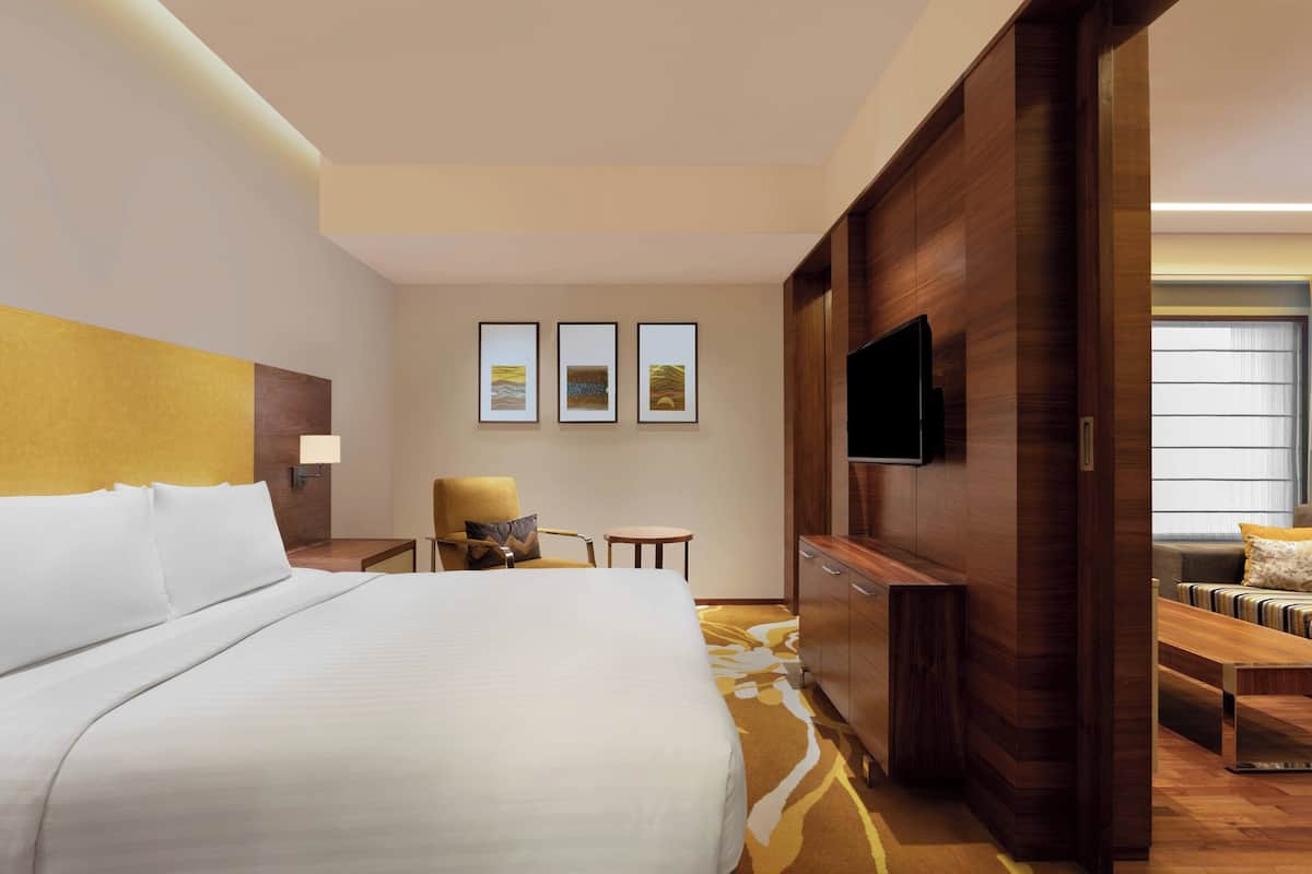 A hotel room with a large bed, a TV mounted on the wall, a small table with a chair, and a sofa with a cushion.