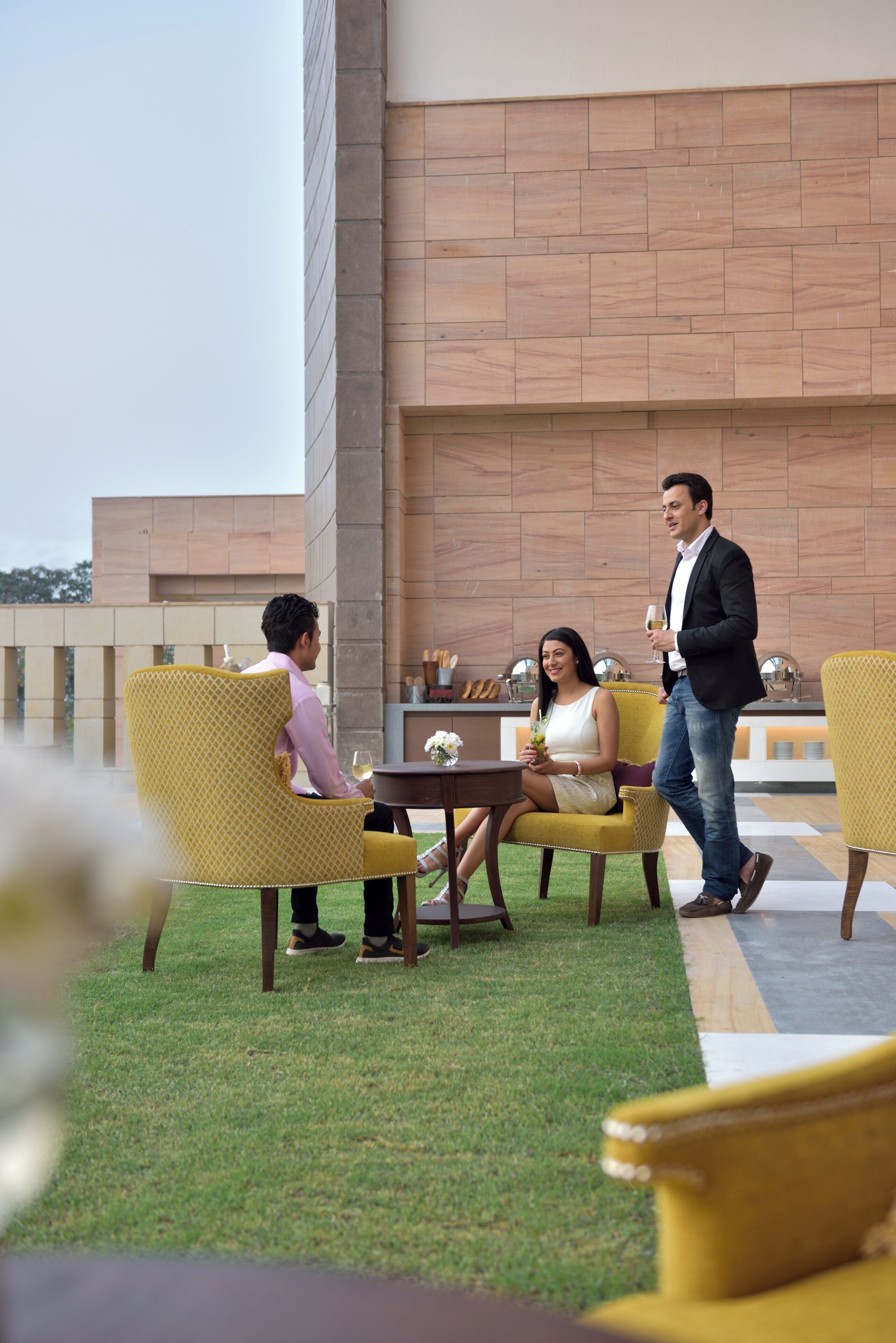 A terrace with yellow chairs and a table, a man and a woman sitting, and a man walking by.