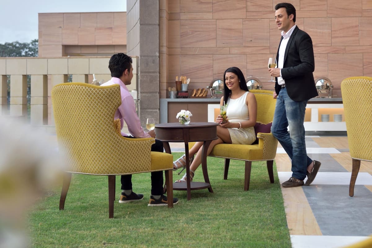 A terrace with yellow chairs and a table, a man and a woman sitting, and a man walking by.