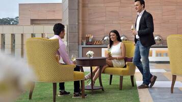 A terrace with yellow chairs and a table, a man and a woman sitting, and a man walking by.