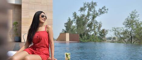 Una persona sentada junto a una piscina, leyendo un libro y tomando algo.