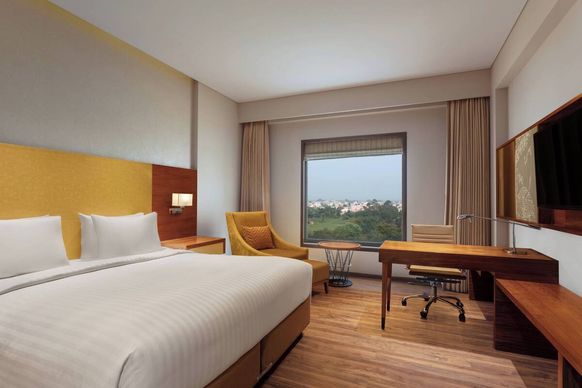 A hotel room with a large bed, a desk, a chair, and a view of a cityscape through the window.
