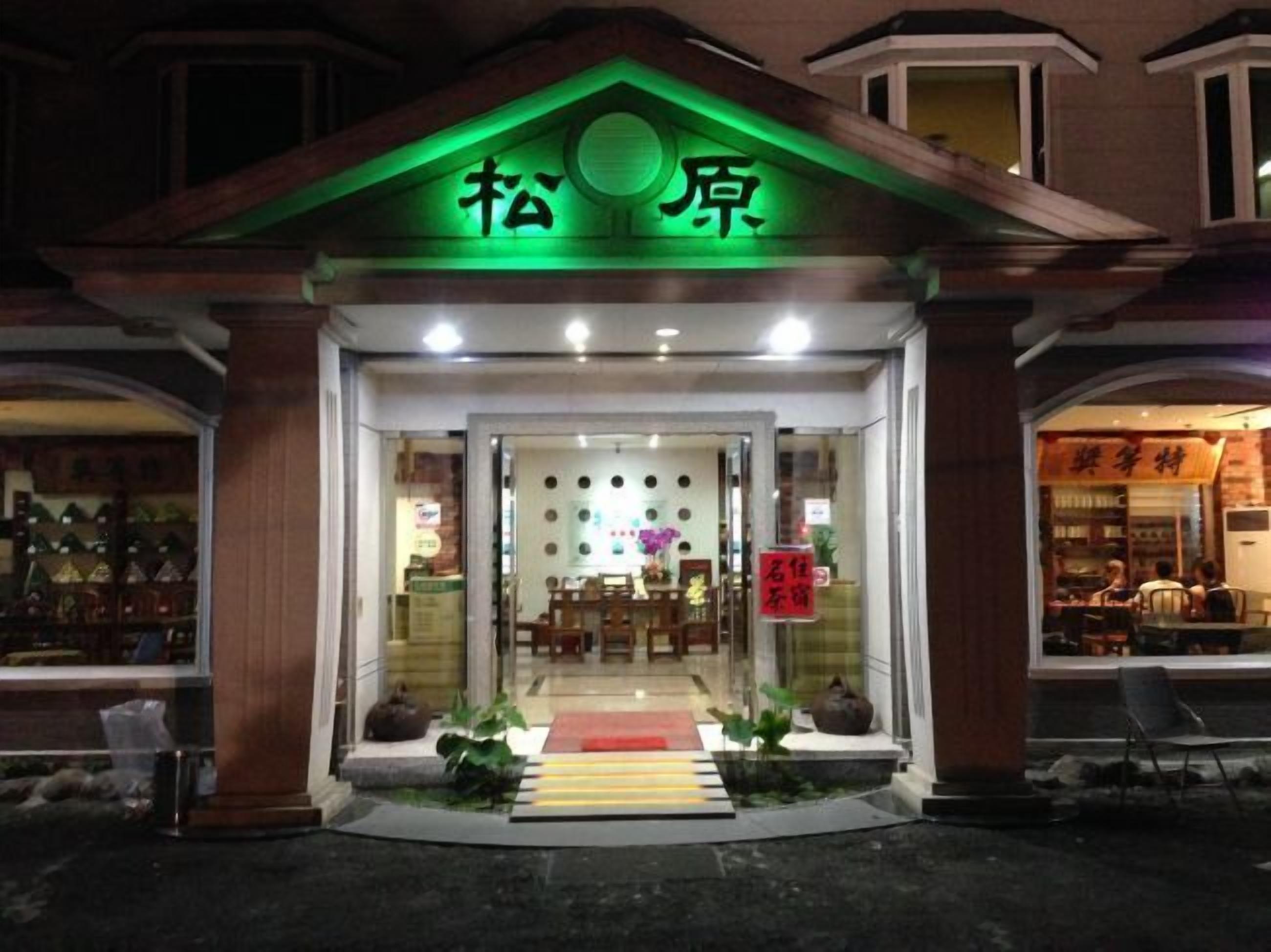 A building with a green-lit entrance and a sign above it. Inside, there are tables and chairs arranged for dining.