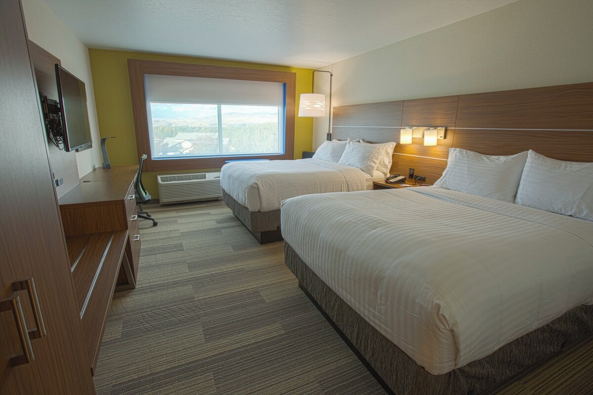 A hotel room with two beds, a desk with a computer, a window with a view, and a lamp.