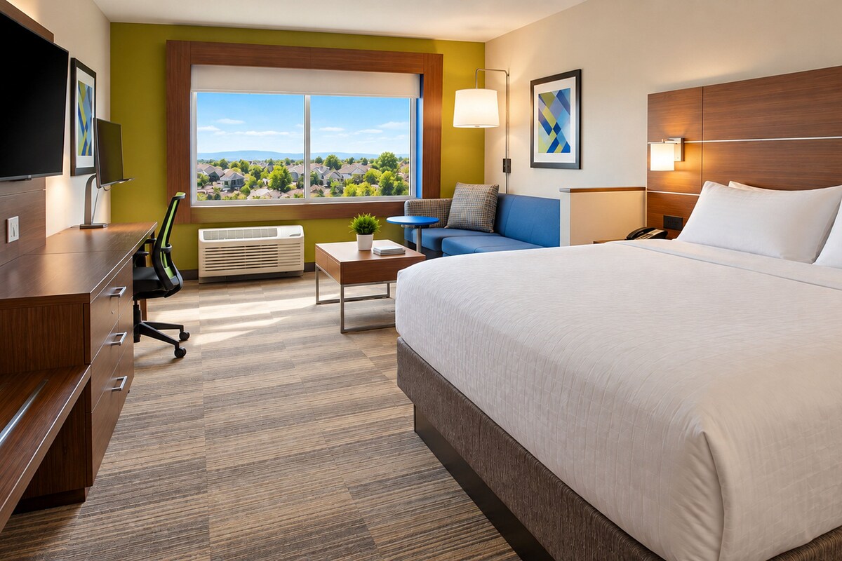 A hotel room with a large bed, a desk, a chair, a small table, a lamp, a TV, and a window with a view of houses.