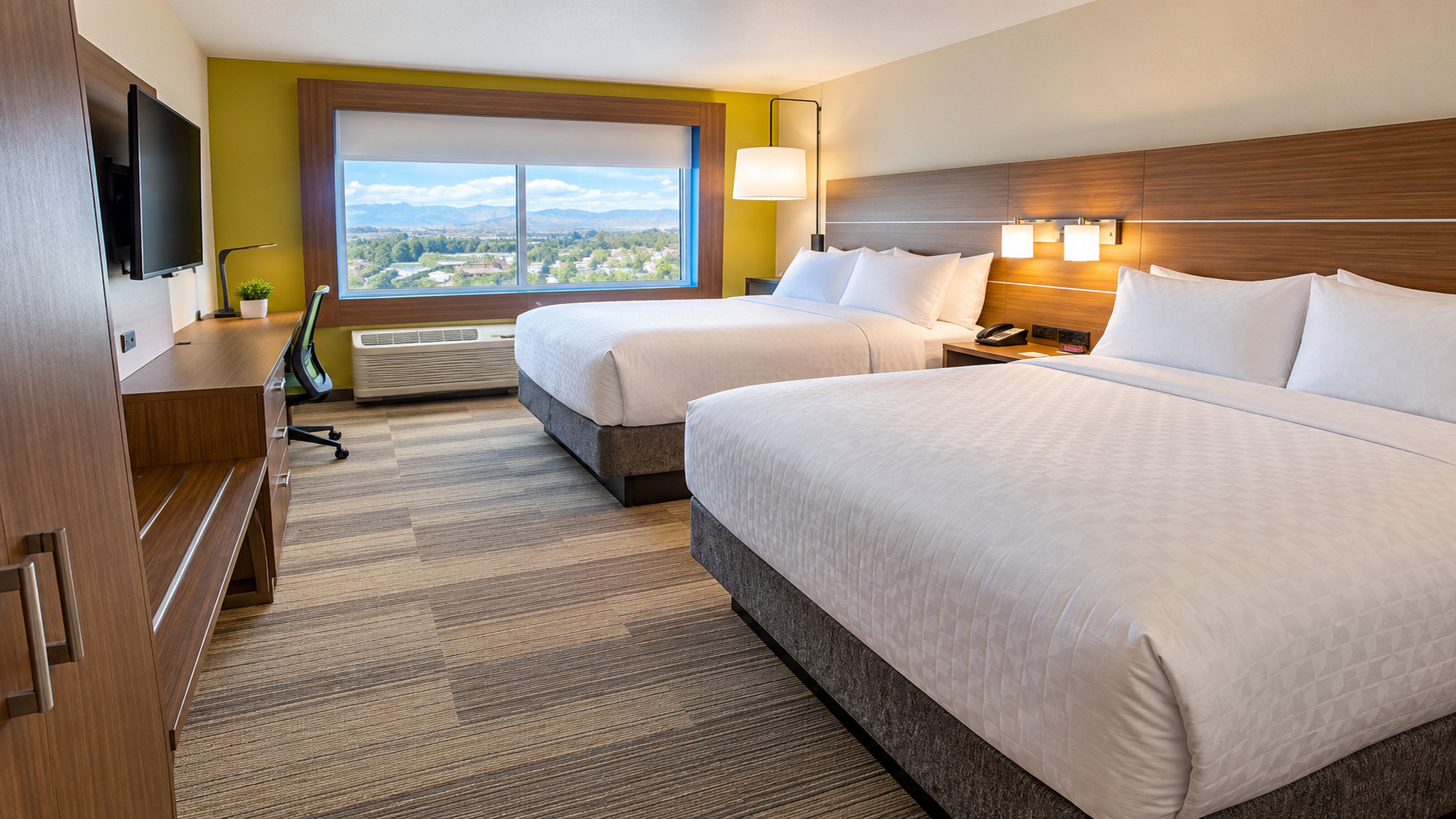 Ein Hotelzimmer mit zwei Betten, einem Flachbildfernseher, einem Schreibtisch und einem großen Fenster mit Blick auf die Stadt.