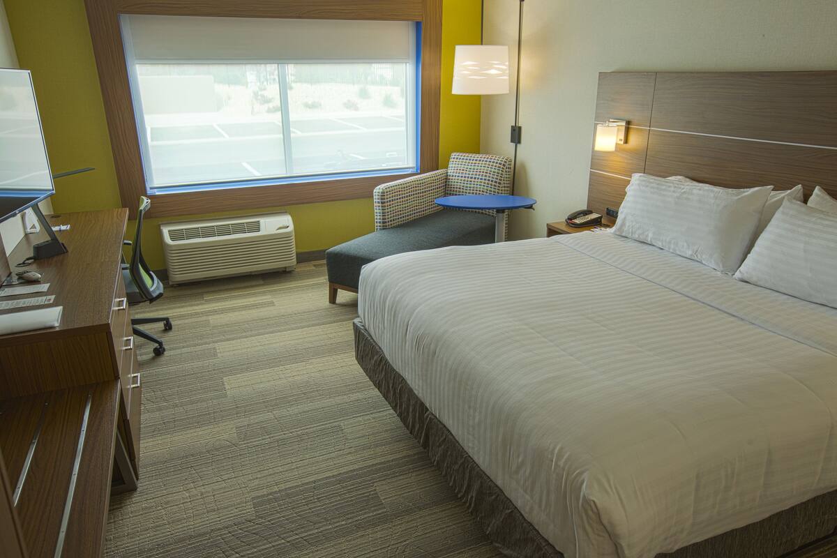 A hotel room with a large bed, a desk with a computer, a chair, a small table, and a window with a view of a parking lot.