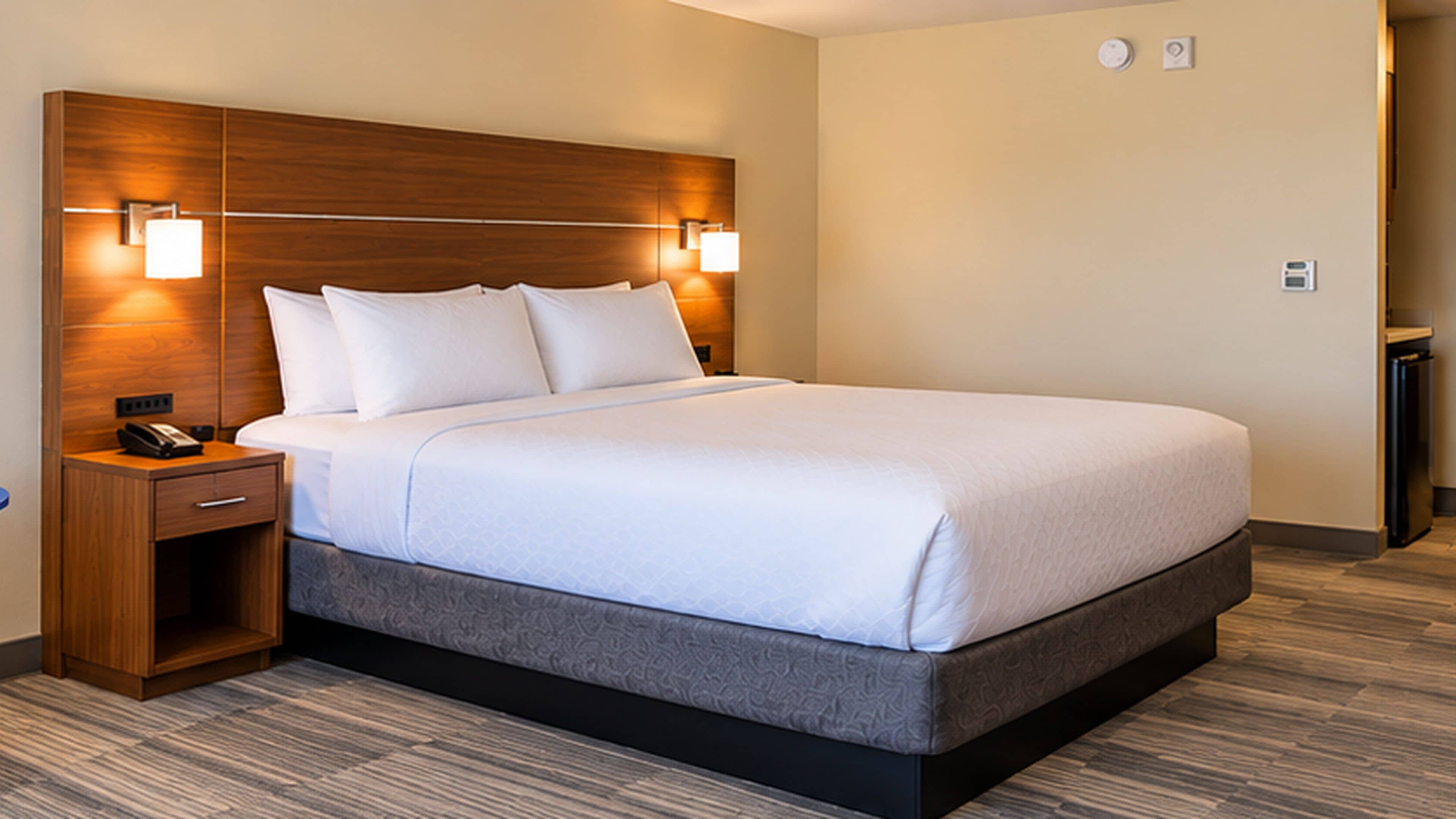 A hotel room with a large bed, a wooden headboard, bedside lamps, a nightstand, and a small drawer.