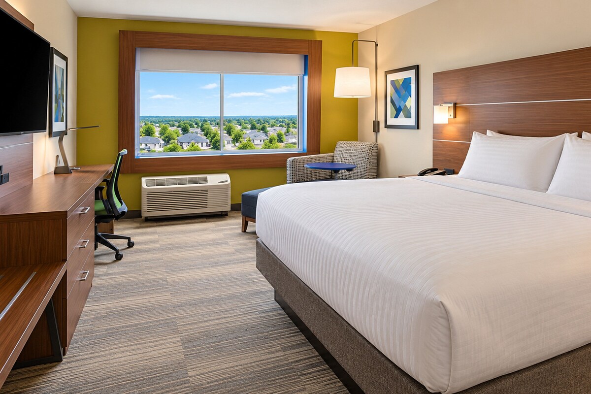 A hotel room with a large bed, a desk, a chair, a window with a view, and a wall-mounted TV.