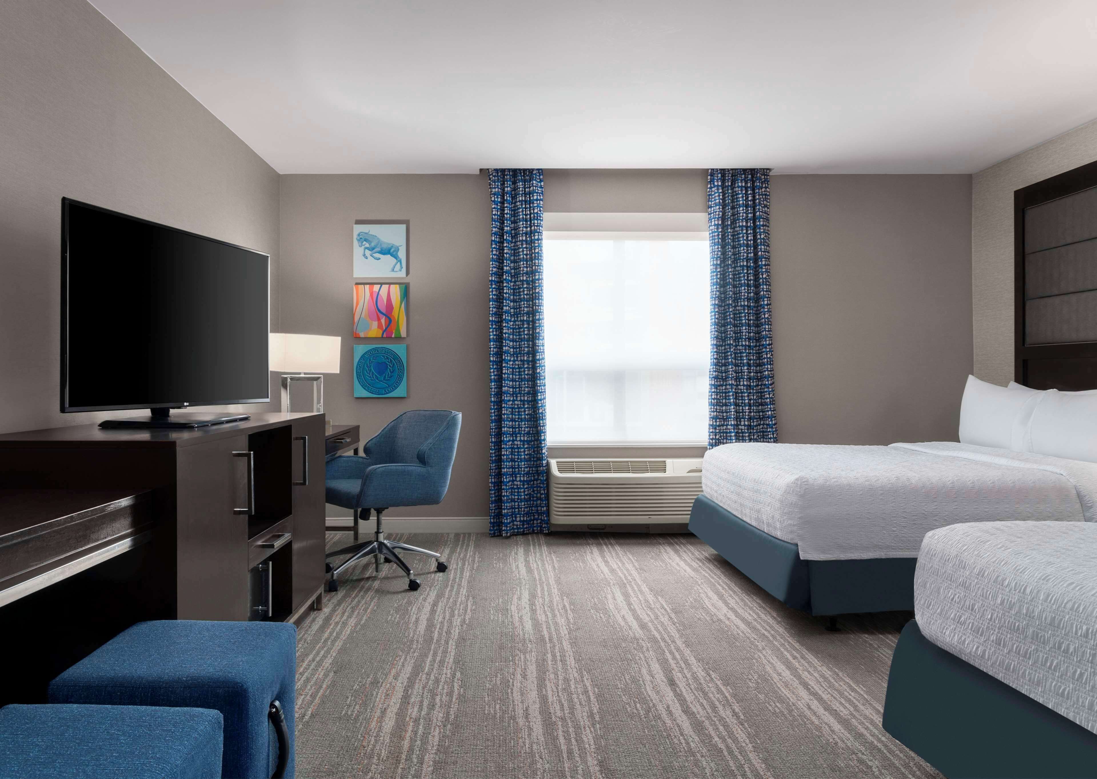 A hotel room with a flat-screen TV, a desk with a lamp, a blue chair, two beds, a window with blue curtains, and a wall-mounted air conditioner.