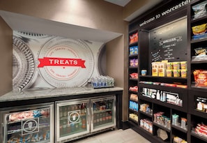 A snack bar with a 'Treats' sign, refrigerated drink dispensers, and shelves stocked with various snacks and beverages. - Hampton Inn & Suites Worcester (Worcester)