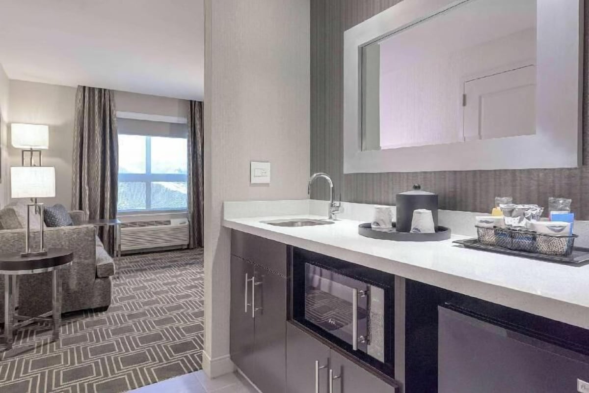 Una habitación de hotel moderna con kitchenette, un ventanal con cortinas, un sofá y una lámpara.