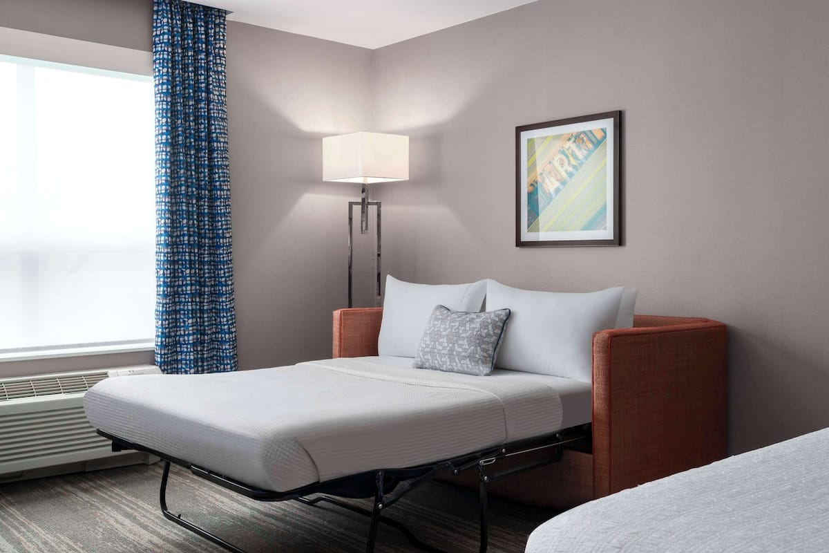 Une chambre d’hôtel avec un lit simple, une lampe sur pied, une œuvre d’art encadrée au mur et une fenêtre avec un rideau bleu.