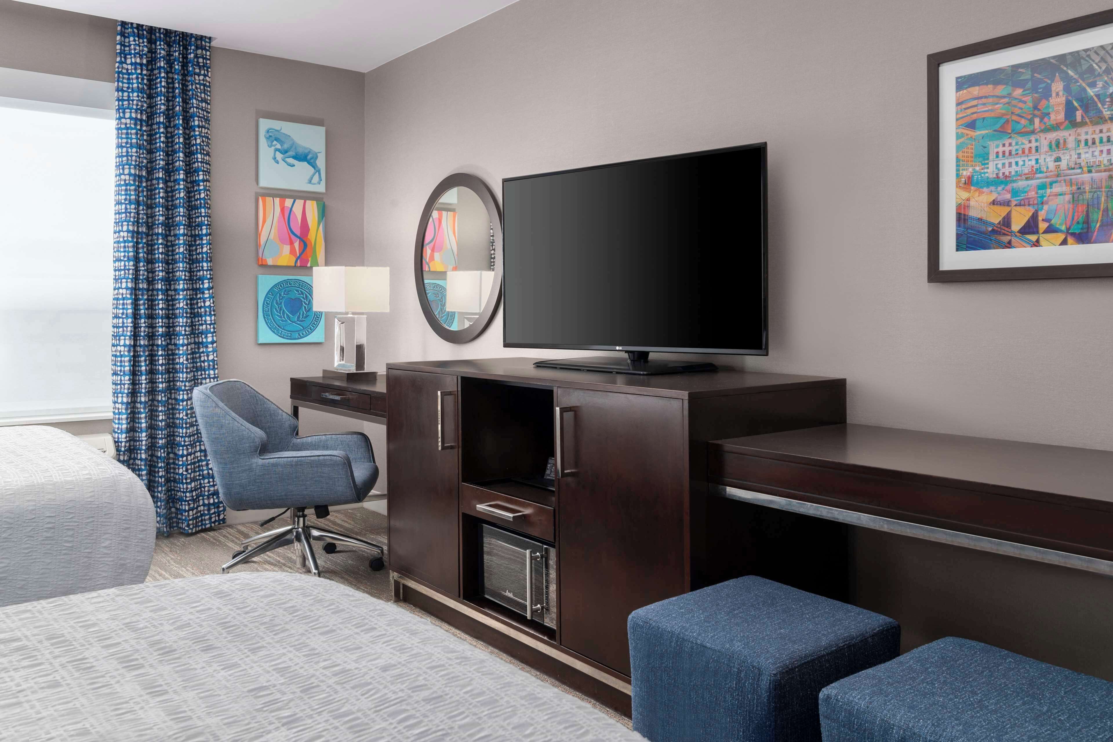 Une chambre d’hôtel comprenant un lit, un bureau avec un téléviseur à écran plat, une chaise, une lampe de bureau et des œuvres d’art accrochées aux murs.