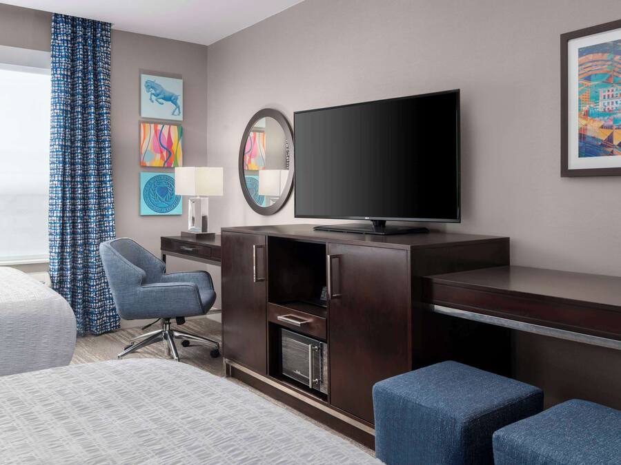 Une chambre d’hôtel comprenant un lit, un bureau avec un téléviseur à écran plat, une chaise, une lampe de bureau et des œuvres d’art accrochées aux murs.