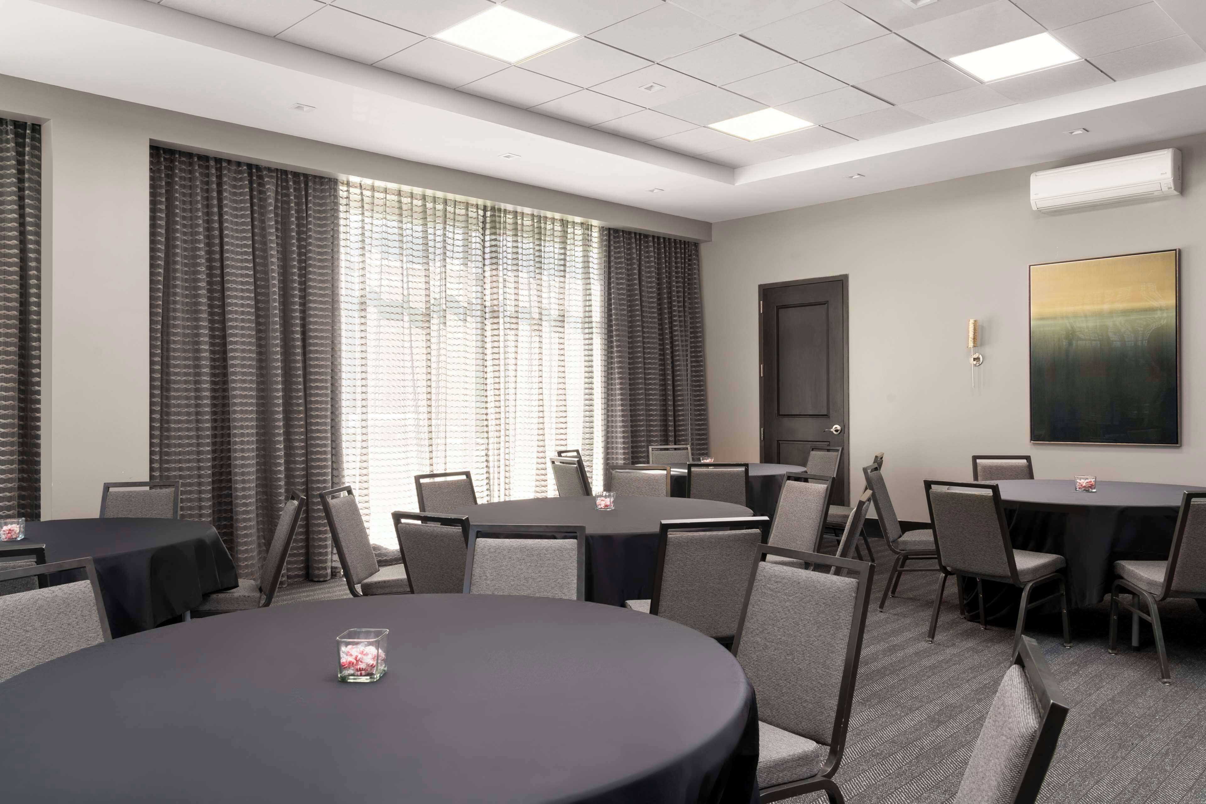 A conference room with round tables and chairs, large windows with curtains, and a painting on the wall.