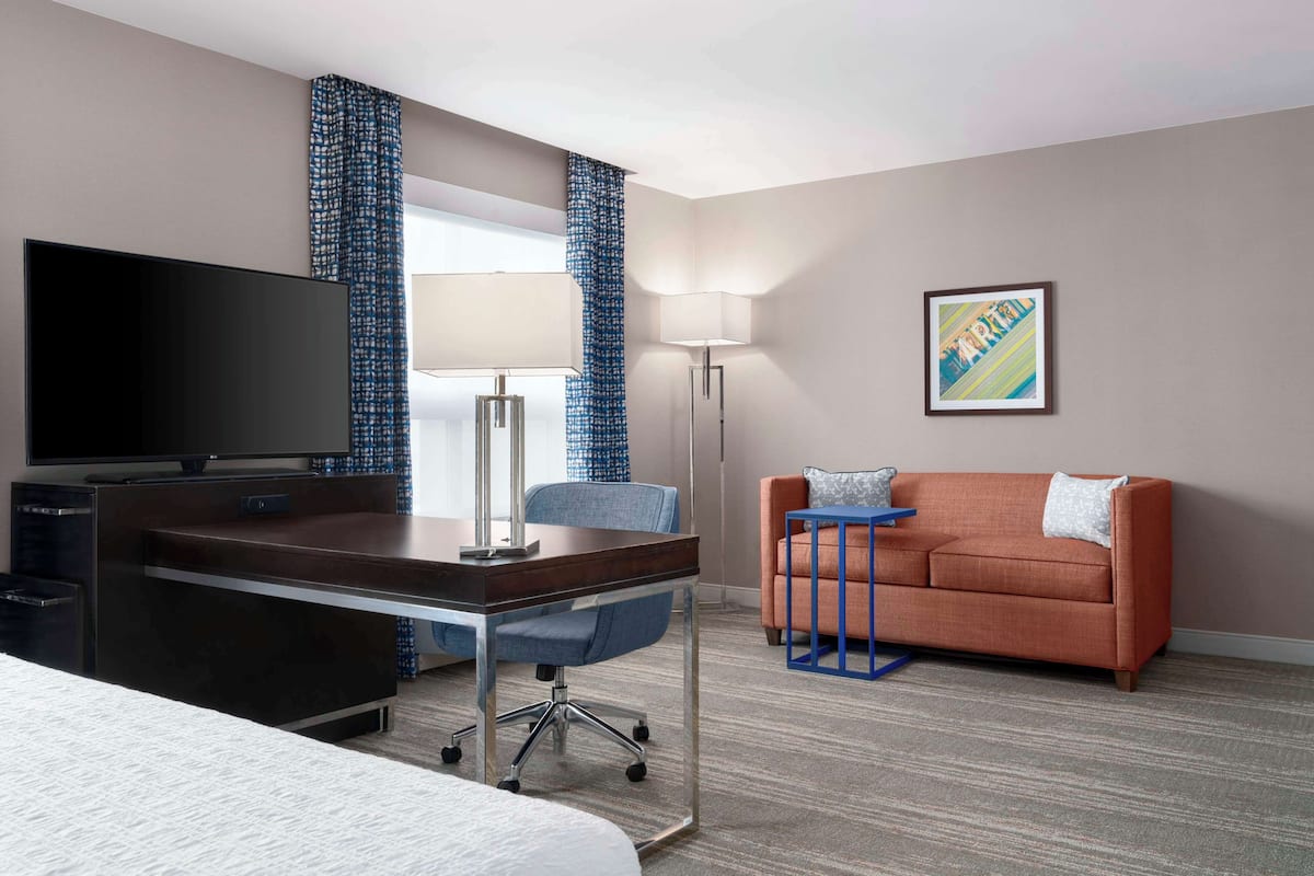 Une chambre d’hôtel comprenant un lit, un bureau avec une télévision, une chaise, une table de chevet bleue, un canapé, une lampe sur pied et un tableau au mur.