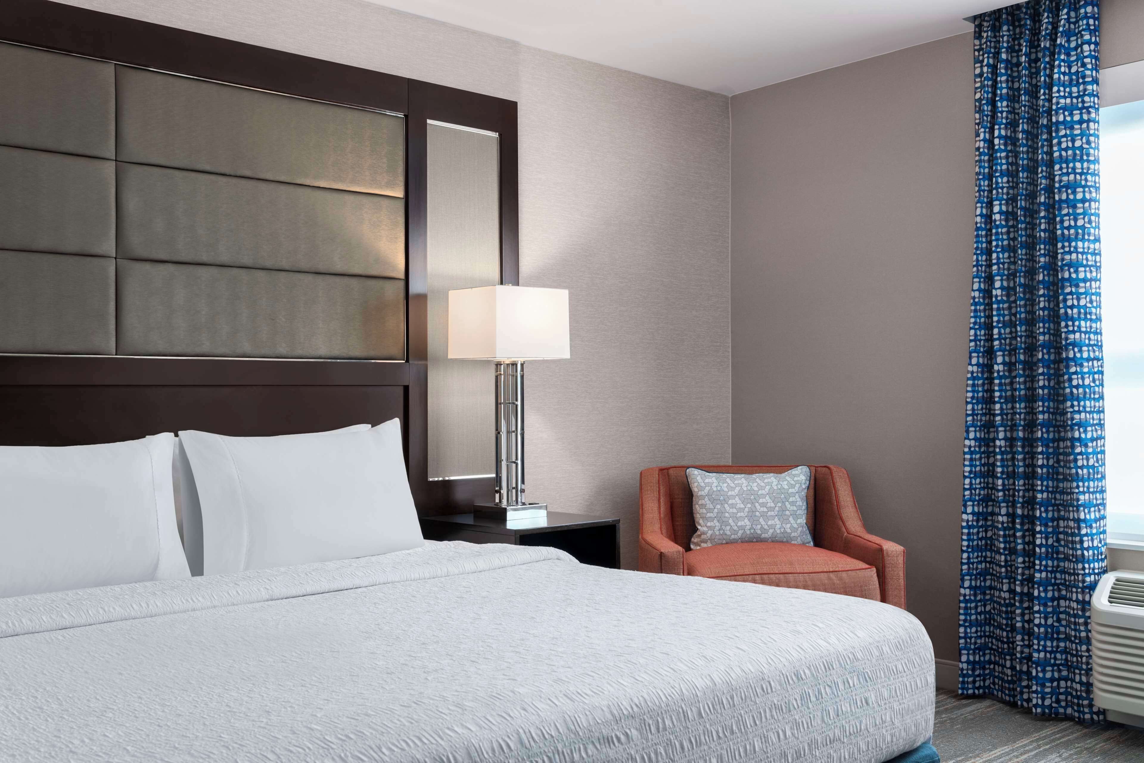 Een hotelkamer met een groot bed, een nachtkastje met lamp, een stoel en een raam met gordijnen.