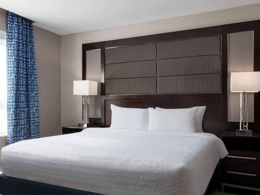 A hotel room with a large bed, dark wood headboard, two bedside tables with lamps, and a blue patterned curtain.