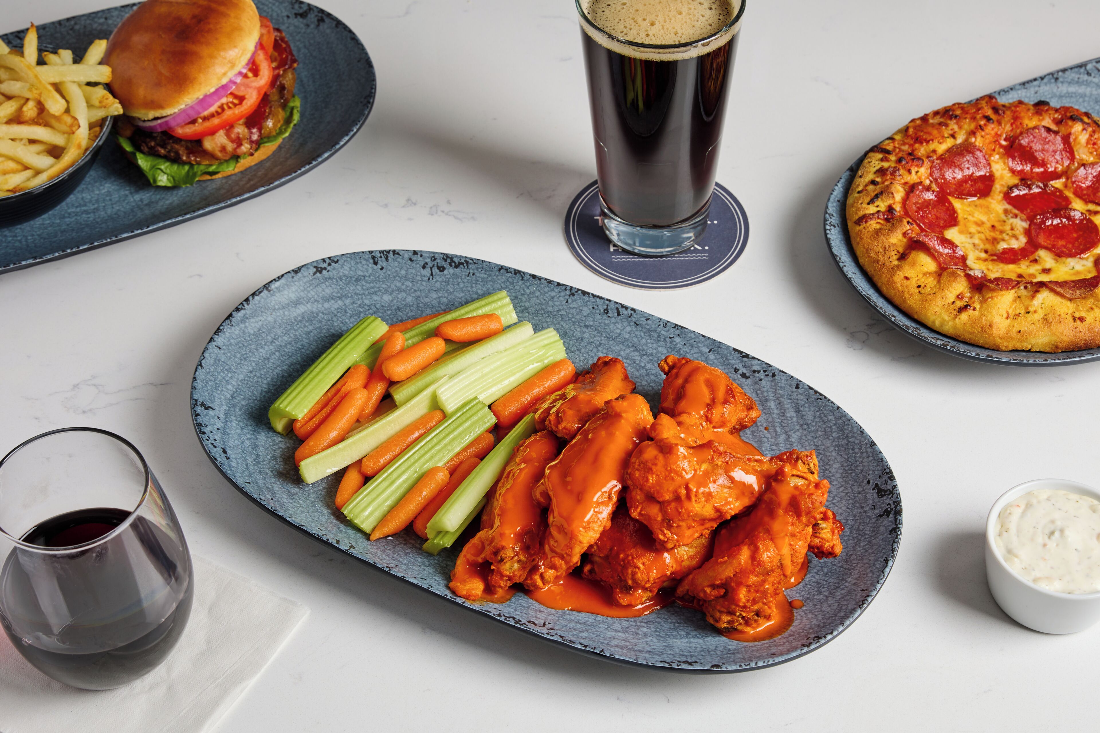 A meal with wings, celery, and carrots on a plate, a burger, fries, a glass of beer, and a pizza.