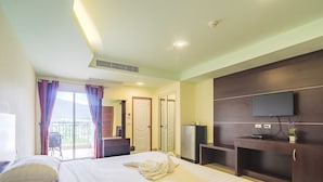 A hotel room with a large bed, a flat-screen TV mounted on the wall, a small desk with a computer, and a balcony with a chair. - Smile Residence (Chalong)