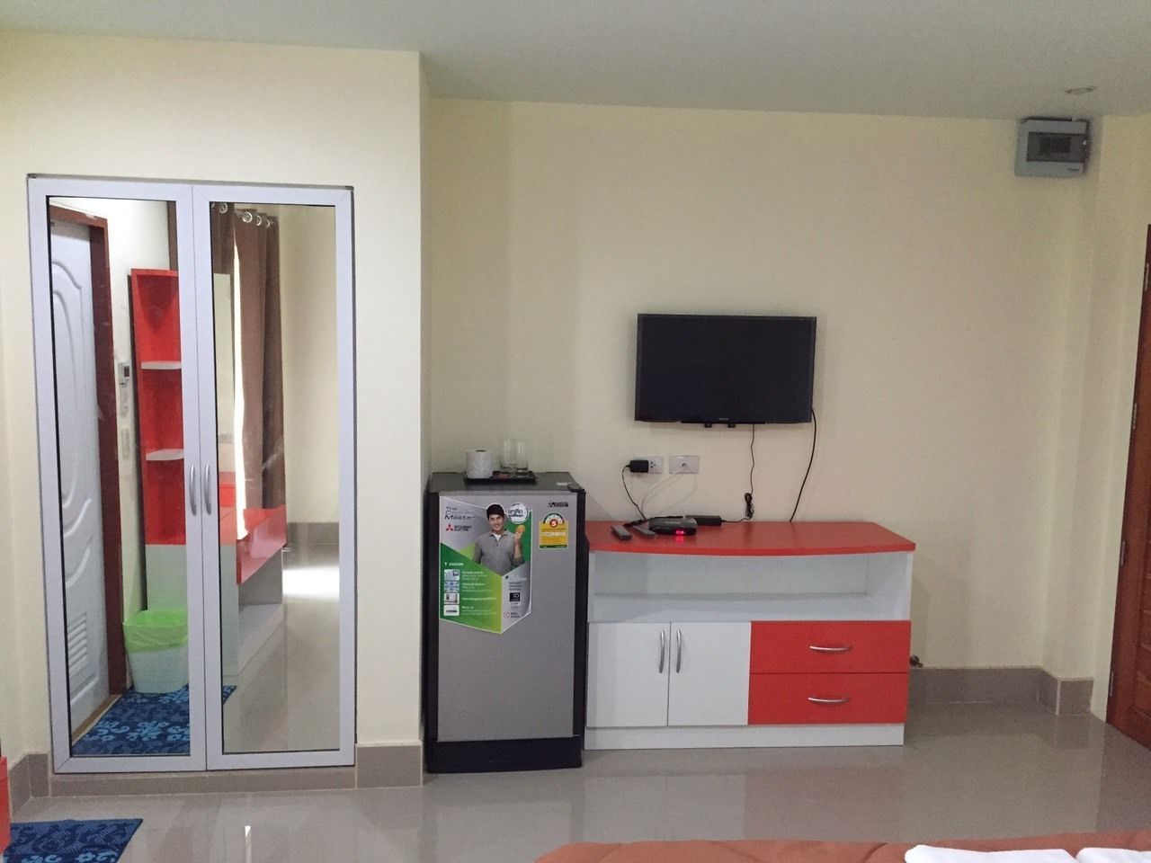 A compact room with a wardrobe, a small refrigerator, a TV mounted on the wall, and a red and white cabinet.