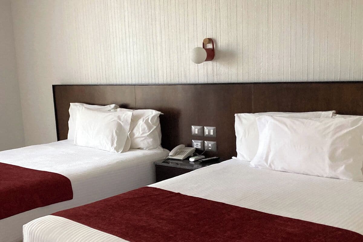 Habitación de hotel con dos camas, cada una con ropa blanca de cama y mantas rojas. Se ve una mesita de noche con un teléfono y una lámpara de pared.