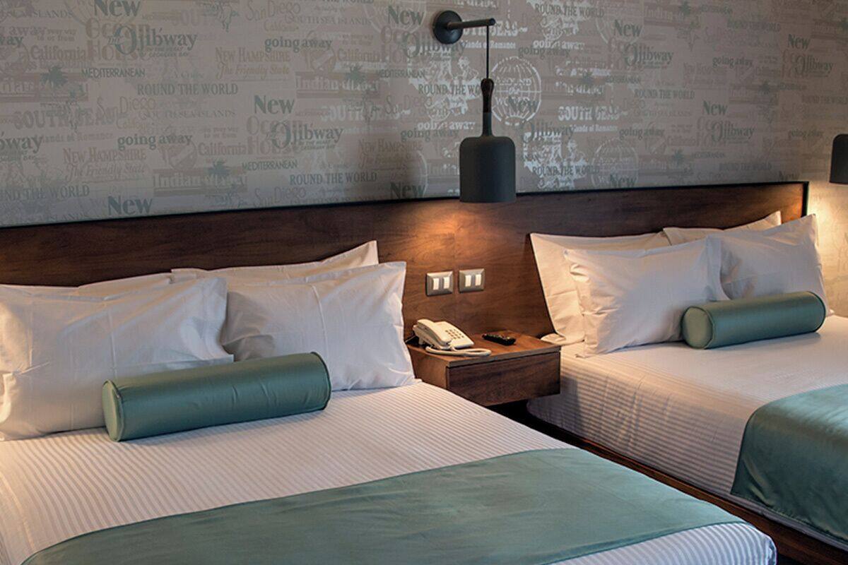 Habitación de hotel con dos camas, cada una con ropa de cama blanca y detalles azul verdosos, cabeceros de madera, mesitas de noche con lámparas y una pared con diseño de mapa del mundo.