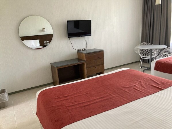 A hotel room with a bed, a TV mounted on the wall, a small wooden dresser, a round mirror, and a small table with chairs. - Hotel Parque Acuático La Caldera (Abasolo)