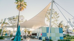 An outdoor dining area with tables and chairs, a large umbrella, and a building with a geometric facade.