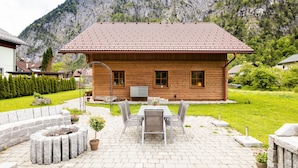 Una casa in legno con tetto in tegole, un patio con tavolo e sedie, e un giardino con una panchina in pietra.