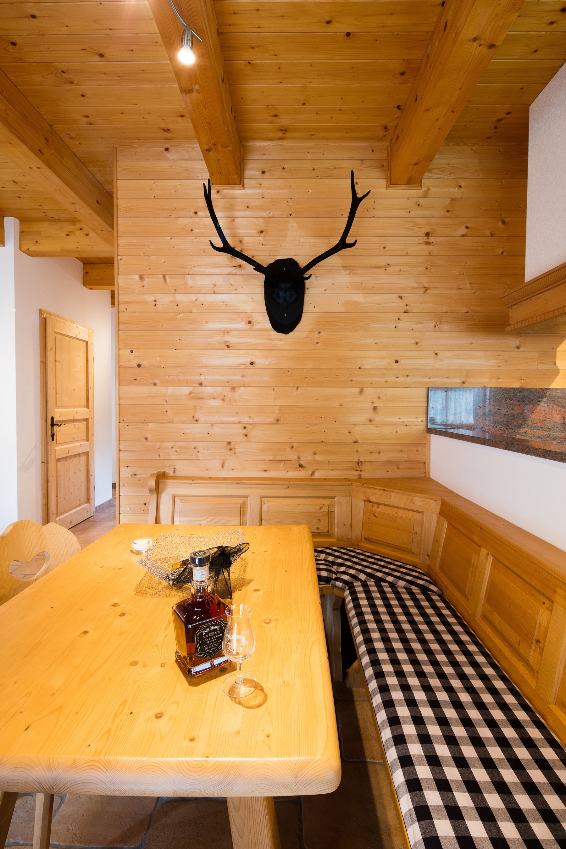 Ein Holzinterieur mit einer karierten Bank, einem Tisch mit Flasche und Glas sowie einem aufgehängten Hirschkopf an der Wand.