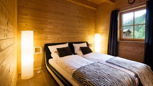 Camera da letto in una baita di legno, con un letto, due lampade sui comodini e una finestra con tende.