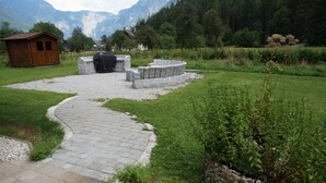 A stone patio with a curved stone bench and a covered grill area, surrounded by a grassy field and a wooden cabin. - W&S Executive Apartments - Hallstatt II (Hallstatt)