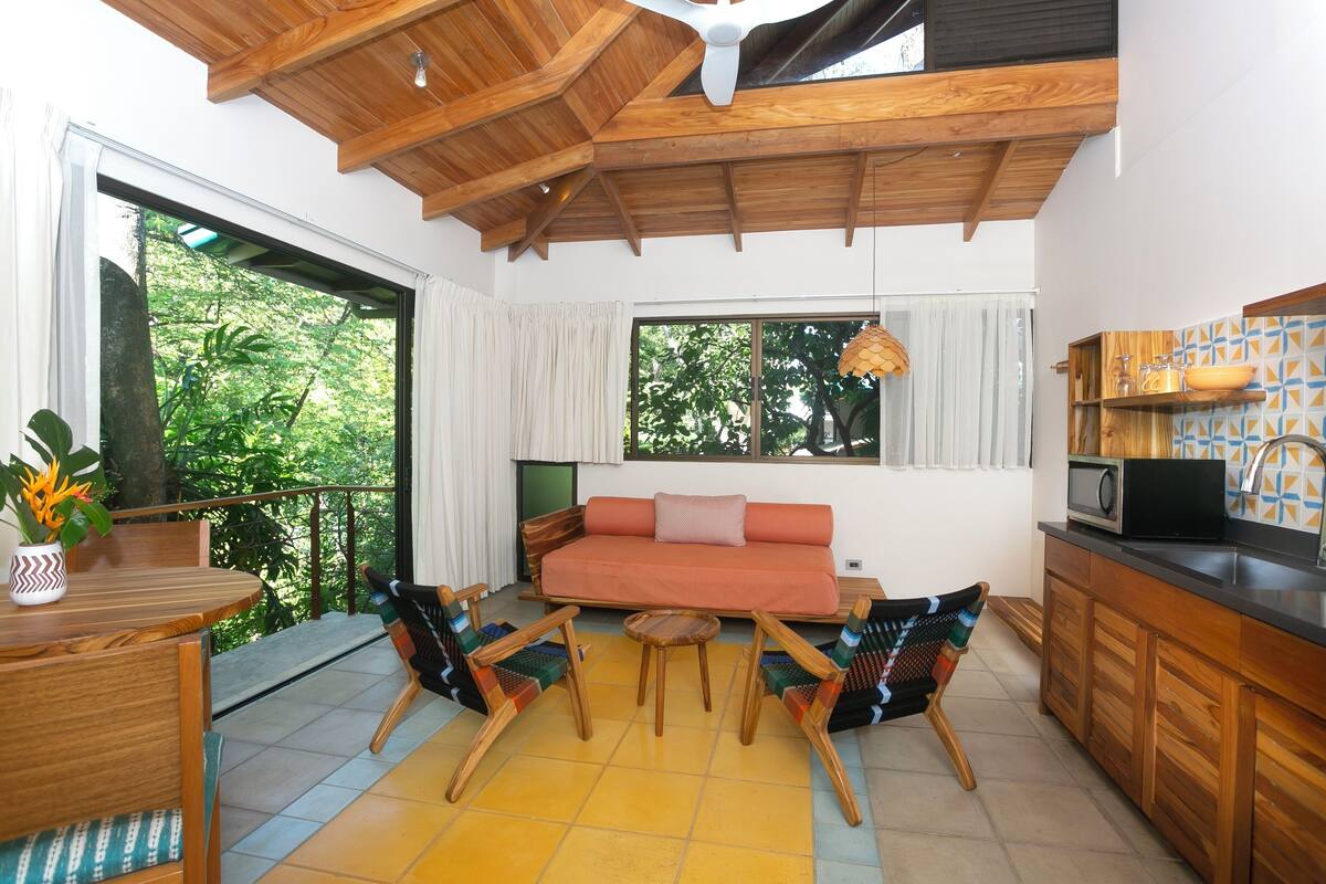 A spacious living area with a wooden ceiling, a sofa, two chairs, a small round table, and a kitchen area with a microwave.