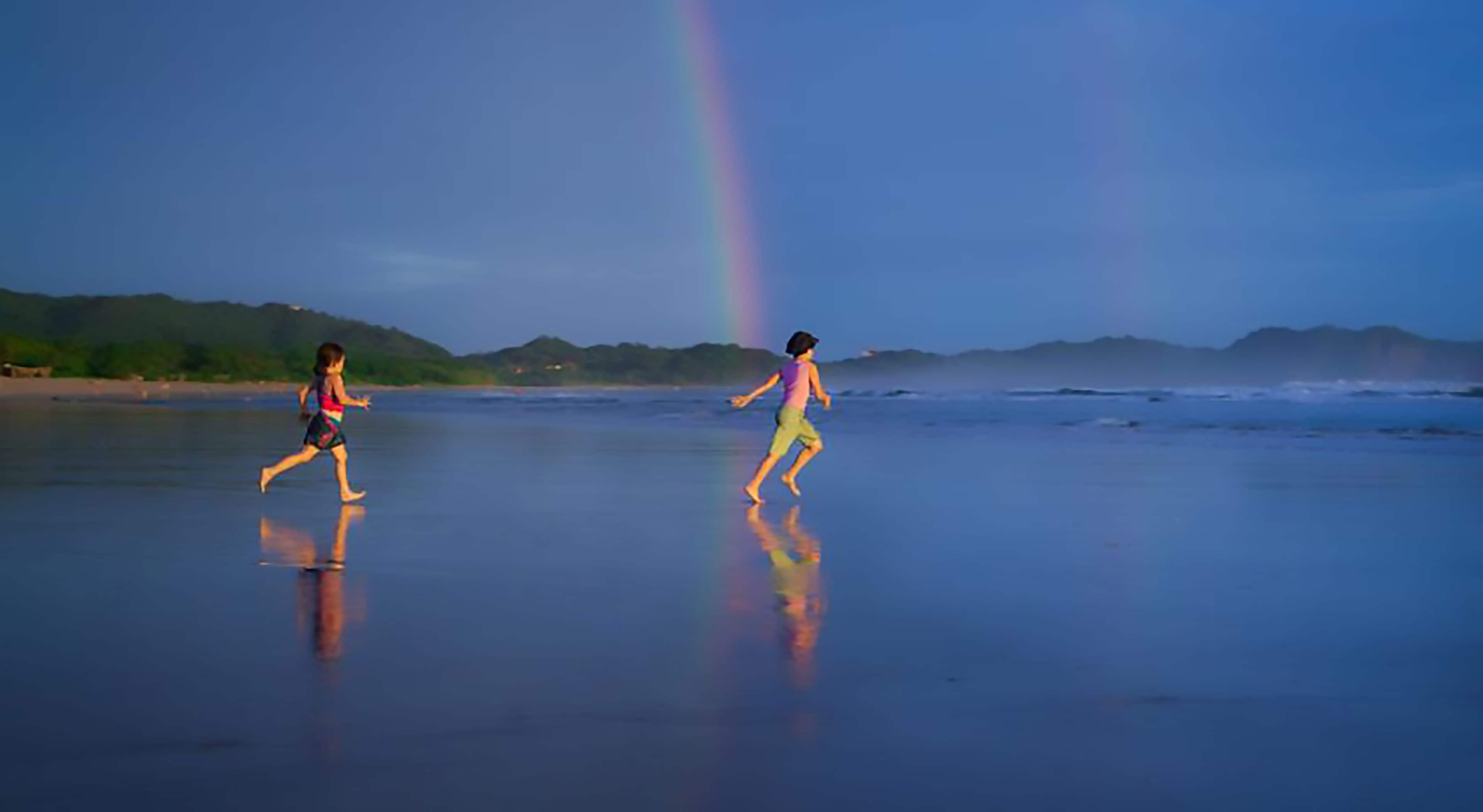 Two children running on a beach with a rainbow in the background.