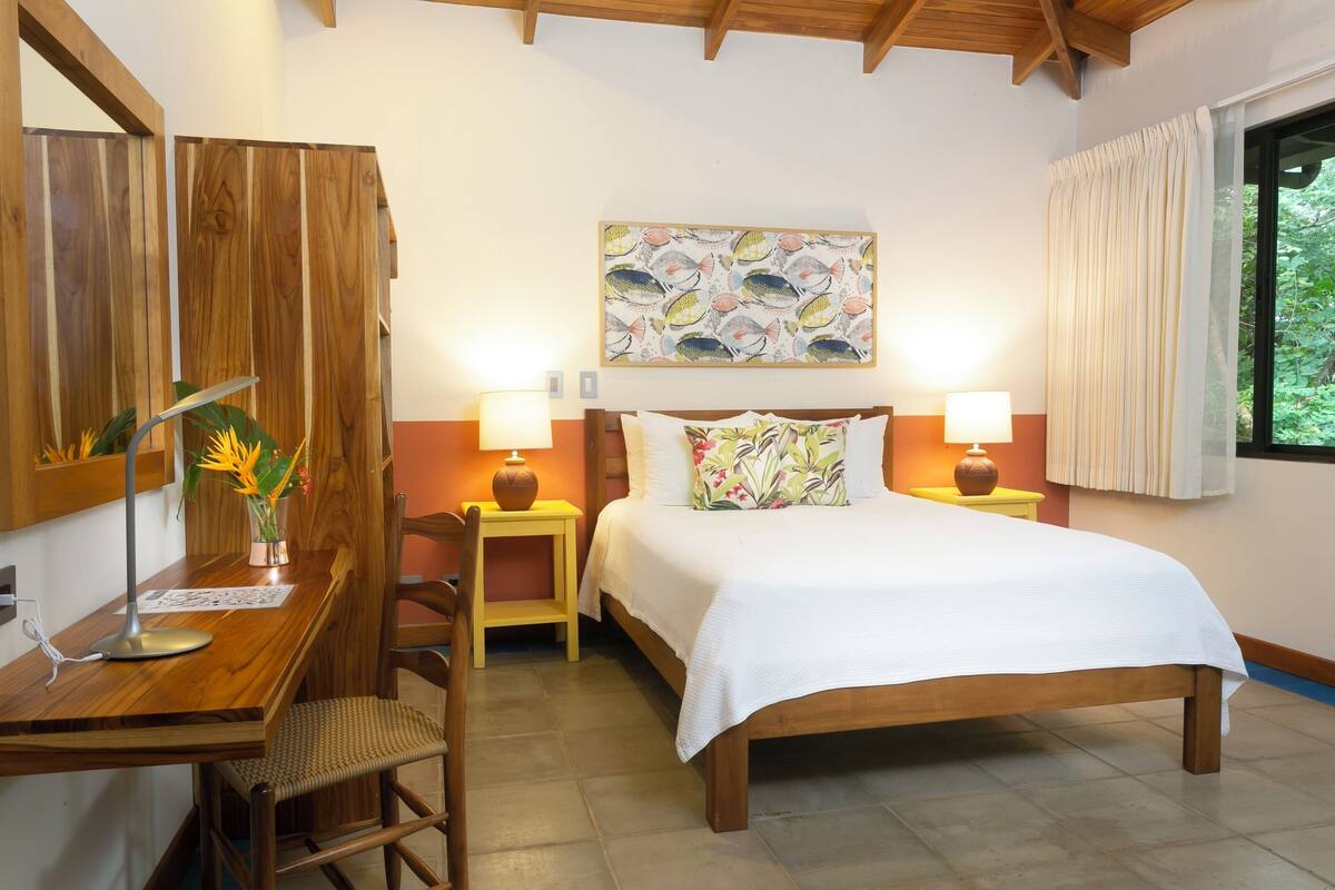 A bedroom with a wooden bed, two bedside tables with lamps, a desk with a chair, a large window with curtains, and a wall-mounted artwork.