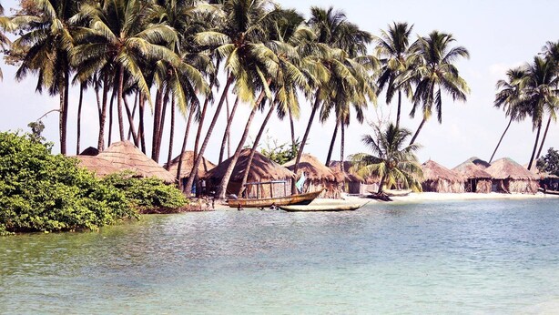 Une plage avec des huttes aux toits de chaume, des palmiers et un bateau sur l’eau.