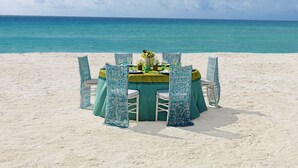 Une table ronde, dressée pour quatre personnes, est installée sur une plage de sable, avec vue sur l’océan.