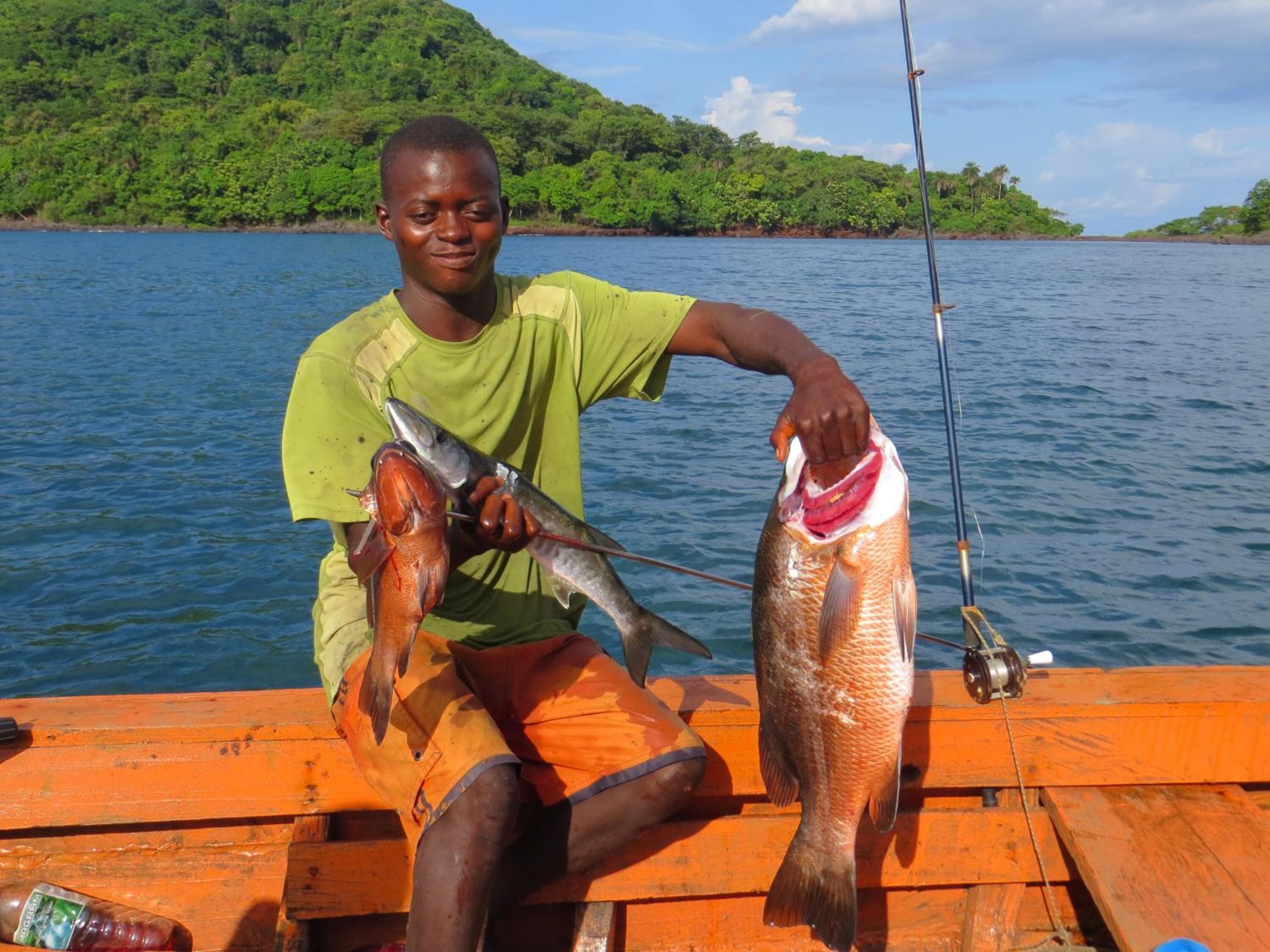 A person holding two fish on a wooden dock with a fishing rod, surrounded by water and greenery.