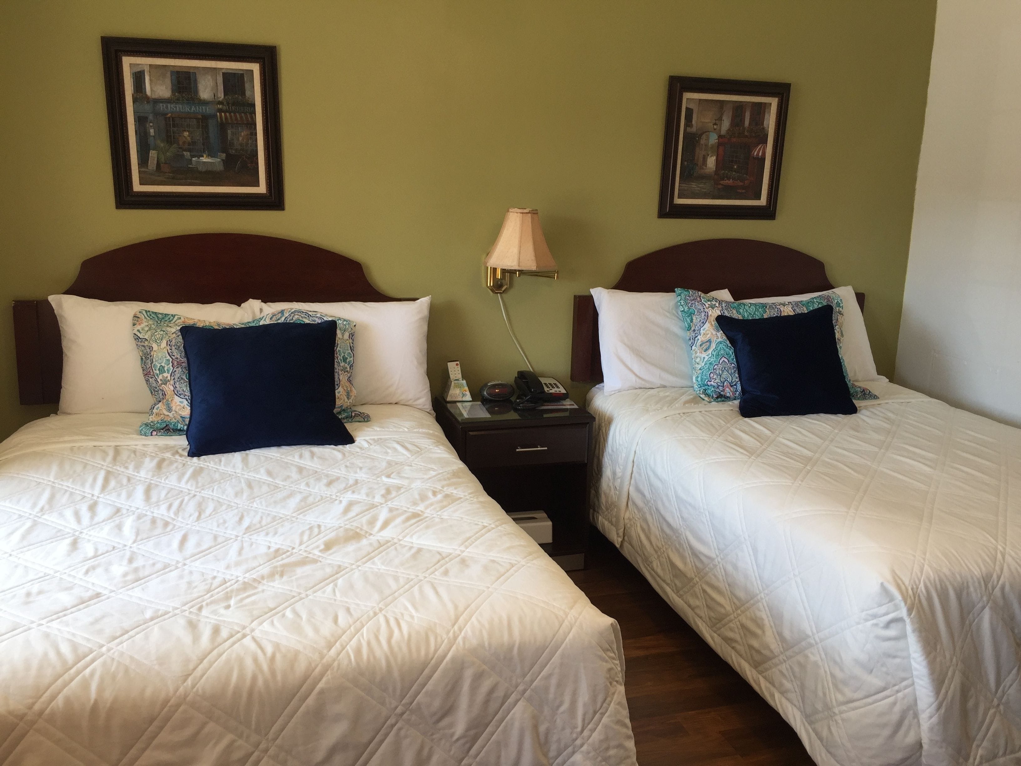Two queen beds with white linens and patterned pillows, a bedside table with a lamp, and two framed pictures on the walls.