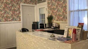A reception desk with a patterned wallpaper, a chandelier, and a window with sheer curtains. - The Baxa Inn (Buckhannon)