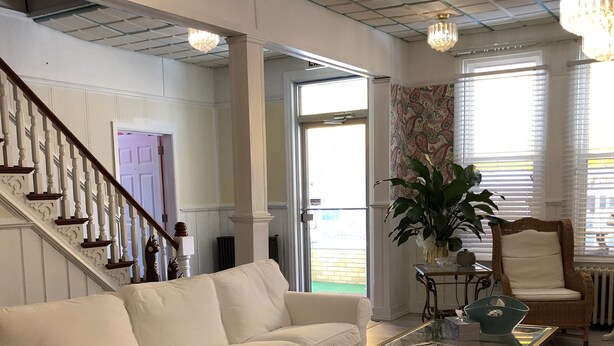 A spacious living room with a white sectional sofa, a glass coffee table, a wicker chair, and a large potted plant. - The Baxa Inn (Buckhannon)