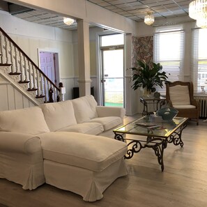 A spacious living room with a white sectional sofa, a glass coffee table, a wicker chair, and a large potted plant. - The Baxa Inn (Buckhannon)