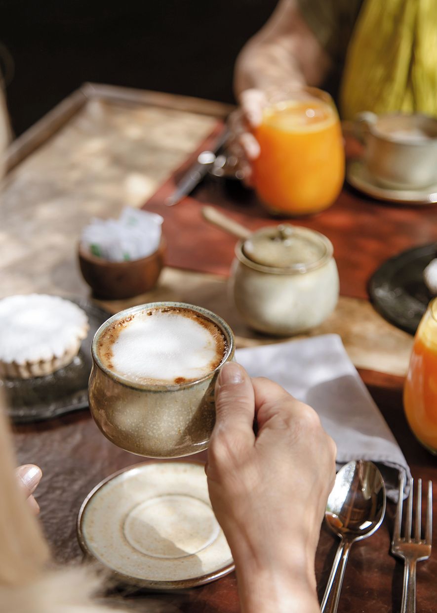 Una mano sostiene una taza de café con espuma, un vaso de zumo de naranja y un cuenco pequeño de azúcar sobre una mesa de madera.