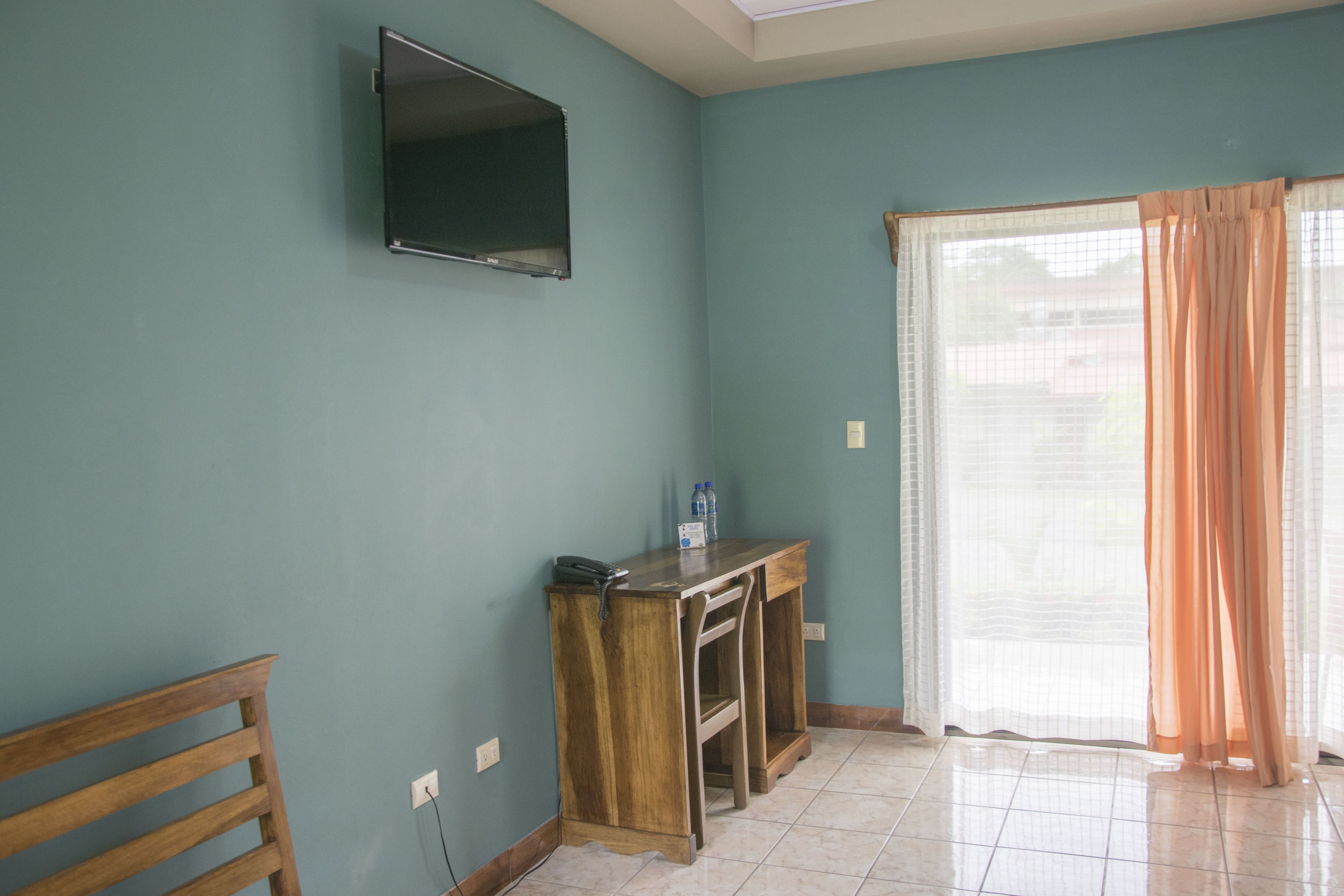 A room with teal walls, a wooden desk with a phone and water bottles, a TV mounted on the wall, and a sliding glass door with curtains.